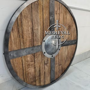 Puede incluir: Un escudo redondo de madera con borde y barras transversales de metal. El escudo presenta un patr&oacute;n de vetas de madera marr&oacute;n claro y un jefe de metal plateado. El texto "MEDIEVAL RELIC" est&aacute; impreso en el escudo.
