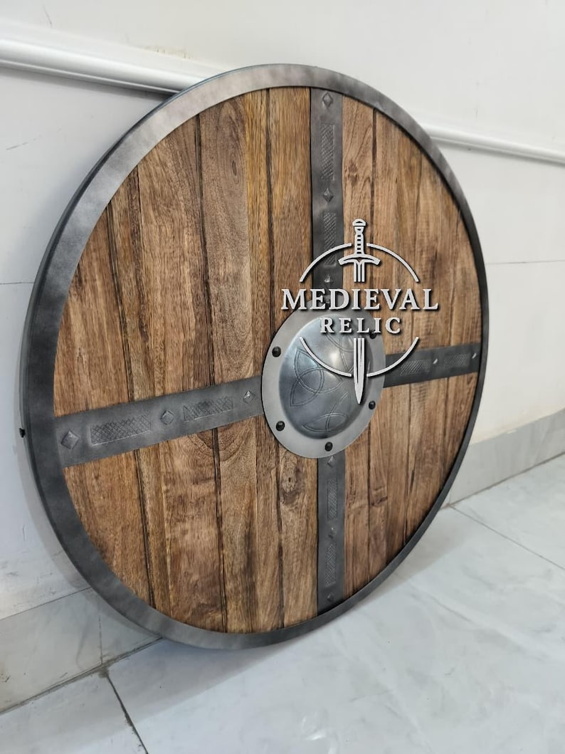 Puede incluir: Un escudo redondo de madera con borde y barras transversales de metal. El escudo presenta un patr&oacute;n de vetas de madera marr&oacute;n claro y un jefe de metal plateado. El texto "MEDIEVAL RELIC" est&aacute; impreso en el escudo.