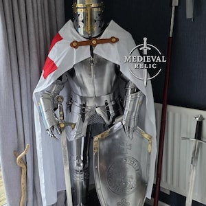 May include: A full suit of medieval knight armor, including a helmet, breastplate, and leg guards. The armor is silver with gold accents and a white cape with a red cross. A shield and sword are also present.