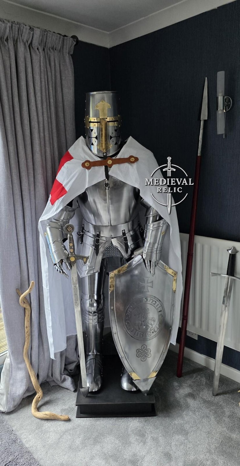 May include: A full suit of medieval knight armor, including a helmet, breastplate, and leg guards. The armor is silver with gold accents and a white cape with a red cross. A shield and sword are also present.