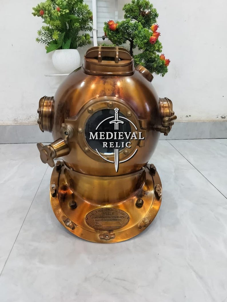 May include: A vintage brass diving helmet with a round, porthole-like window displaying the text "MEDIEVAL RELIC" and a sword emblem. The helmet has a circular base and multiple fittings.