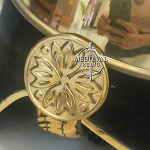 Op de afbeelding: Close-up van een messing helm met een decoratief bloemenembleem. De helm heeft een zwarte fluwelen basis, gouden afwerking en een geweven koorddetail. Het Medieval Relic-logo is zichtbaar.