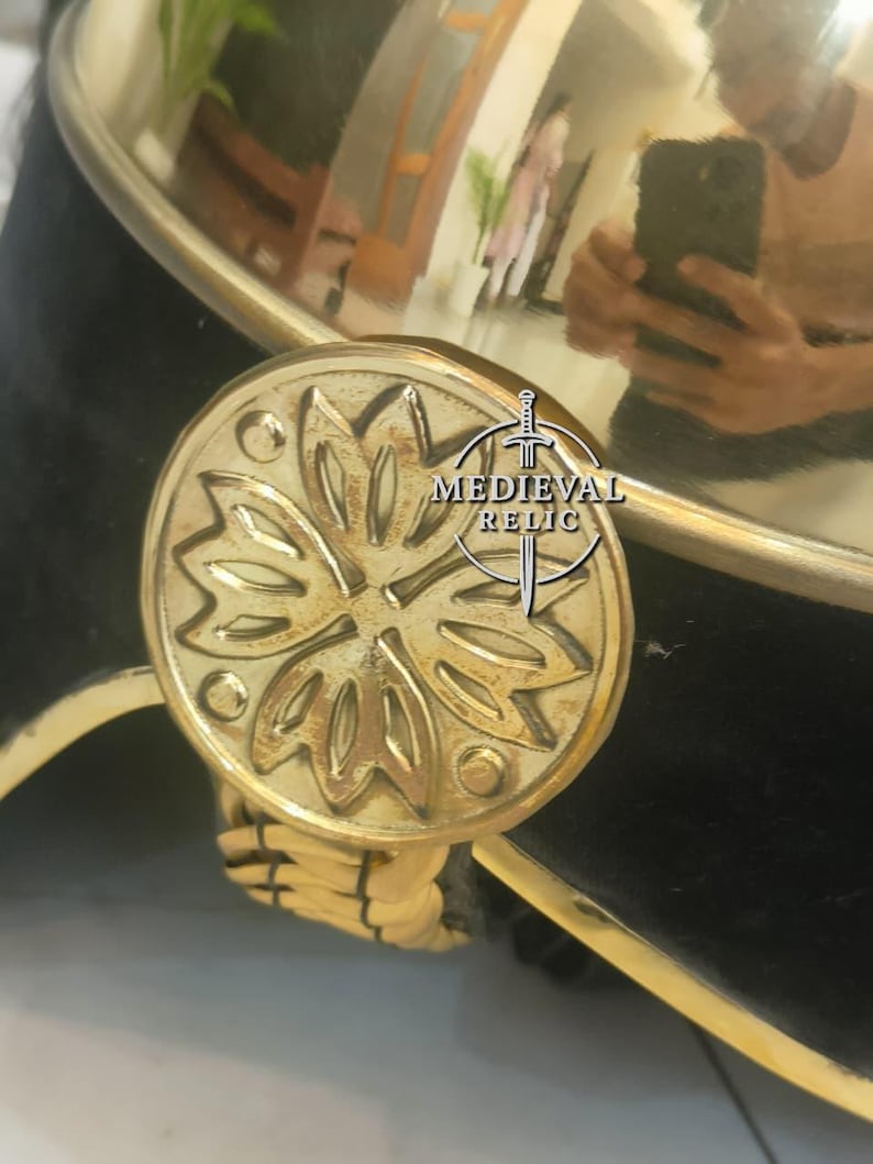 Op de afbeelding: Close-up van een messing helm met een decoratief bloemenembleem. De helm heeft een zwarte fluwelen basis, gouden afwerking en een geweven koorddetail. Het Medieval Relic-logo is zichtbaar.
