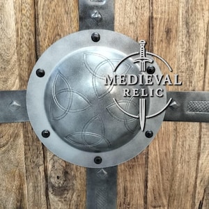 Puede incluir: Un escudo de estilo medieval con fondo de madera y detalles met&aacute;licos. El escudo presenta un centro circular de metal con un dise&ntilde;o de nudo celta, asegurado con correas y remaches met&aacute;licos. El texto "MEDIEVAL RELIC" es visible.