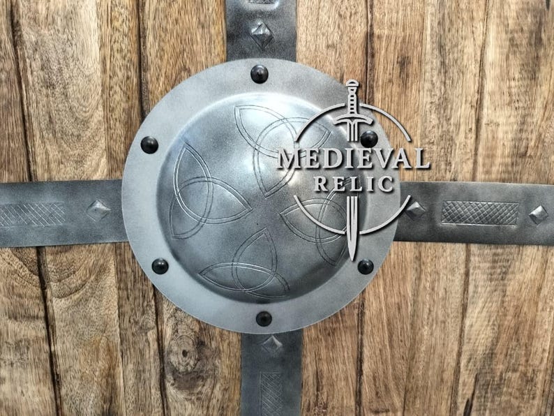 Puede incluir: Un escudo de estilo medieval con fondo de madera y detalles met&aacute;licos. El escudo presenta un centro circular de metal con un dise&ntilde;o de nudo celta, asegurado con correas y remaches met&aacute;licos. El texto "MEDIEVAL RELIC" es visible.