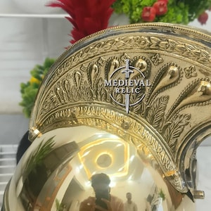 Op de afbeelding: Een gepolijste messing helm met ingewikkelde reli&euml;fdesigns en een rode pluim. De helm draagt de tekst "MEDIEVAL RELIC" en weerspiegelt de omgeving. Een decoratief stuk voor geschiedenisliefhebbers.