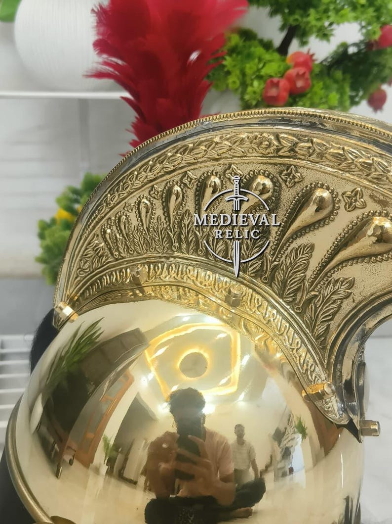 Op de afbeelding: Een gepolijste messing helm met ingewikkelde reli&euml;fdesigns en een rode pluim. De helm draagt de tekst "MEDIEVAL RELIC" en weerspiegelt de omgeving. Een decoratief stuk voor geschiedenisliefhebbers.