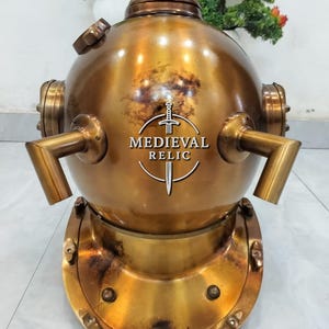 May include: A vintage brass diving helmet with a round top and a wide, bolted base. The helmet has multiple ports and tubes, and the text "MEDIEVAL RELIC" is visible on the front. The brass has a weathered, antique finish.