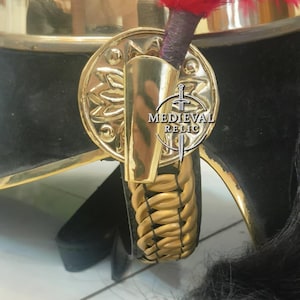 Op de afbeelding: Close-up van een zwart en gouden militaire helm met een rode pluim. De helm heeft een gouden embleem en afwerking, met de tekst "MEDIEVAL RELIC". Een gevlochten gouden riem is ook zichtbaar.