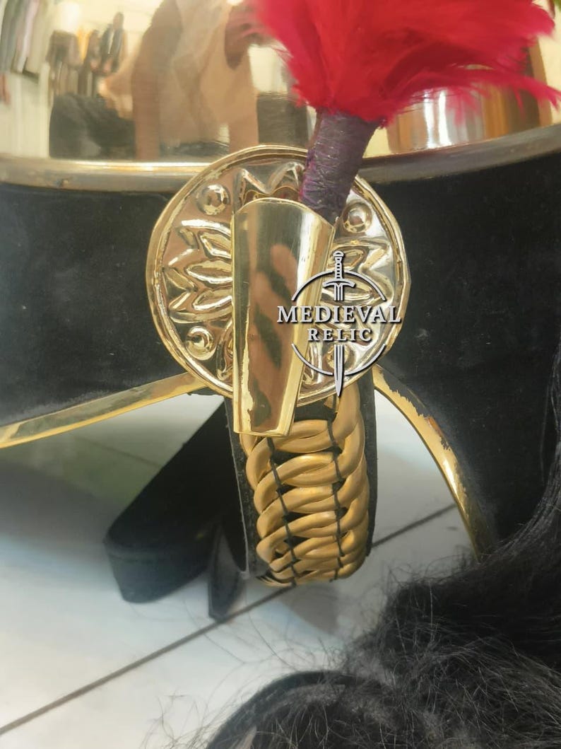 Op de afbeelding: Close-up van een zwart en gouden militaire helm met een rode pluim. De helm heeft een gouden embleem en afwerking, met de tekst "MEDIEVAL RELIC". Een gevlochten gouden riem is ook zichtbaar.