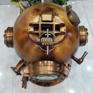May include: A vintage brass diving helmet with a round shape, multiple fittings, and a large front viewing window. The helmet has a weathered, antique appearance with the text "MEDIEVAL RELIC" in a circular logo on the front.