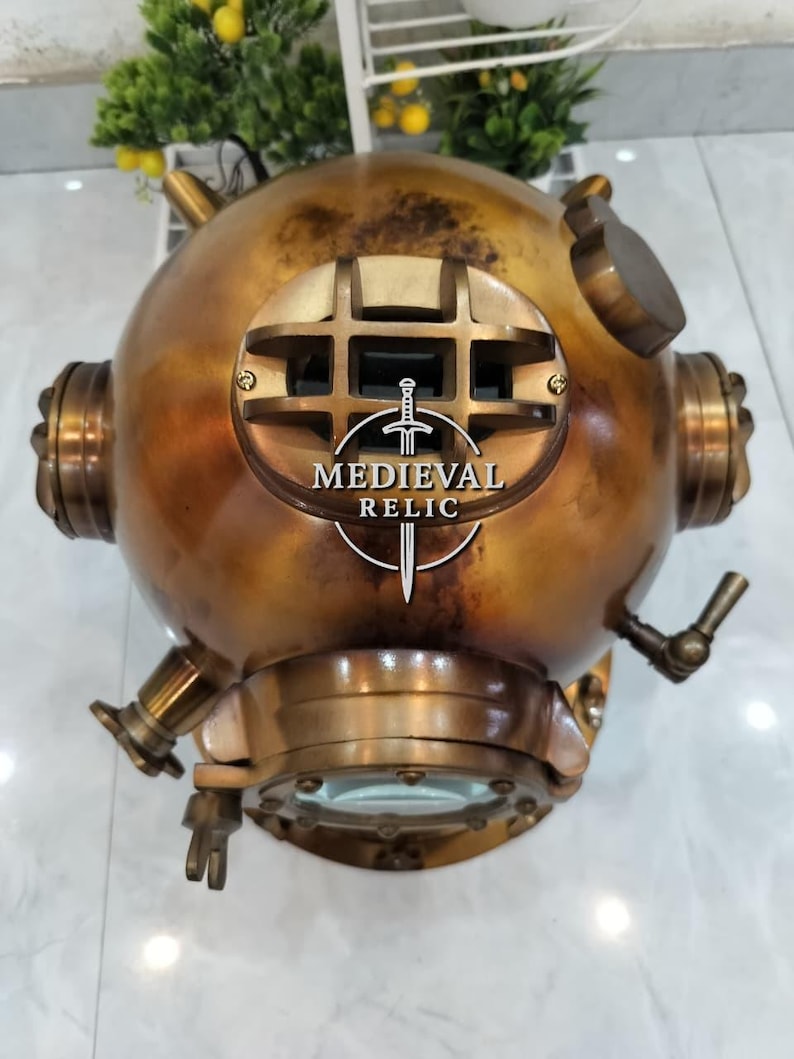 May include: A vintage brass diving helmet with a round shape, multiple fittings, and a large front viewing window. The helmet has a weathered, antique appearance with the text "MEDIEVAL RELIC" in a circular logo on the front.