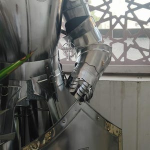 May include: A polished, silver-colored suit of armor with a cross emblem on the chest plate and a gauntlet. The armor is highly reflective, with visible rivets and intricate joint details. The image includes the text "MEDIEVAL RELIC".