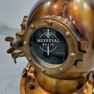 May include: A vintage brass diving helmet with a large circular viewing window. The helmet has a weathered, antique appearance with visible rivets and a plaque that reads "U.S. Navy Diving Helmet". The text "Medieval Relic" is visible on the window.