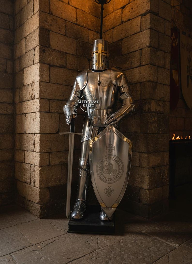 May include: A full suit of polished steel armor, including a helmet, breastplate, and leg guards, stands in a stone room. A sword rests beside the armor, and a shield with a crest is held in place. The armor is from Medieval Relic.