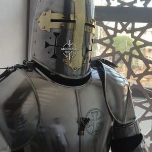 May include: A polished steel knight's helmet with a gold-colored cross design on the front. The helmet is part of a full suit of armor, with a cross emblem on the chest. The image includes the text "MEDIEVAL RELIC".