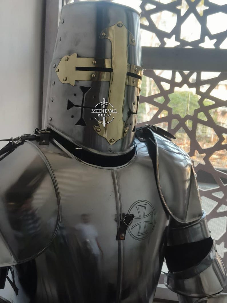 May include: A polished steel knight's helmet with a gold-colored cross design on the front. The helmet is part of a full suit of armor, with a cross emblem on the chest. The image includes the text "MEDIEVAL RELIC".