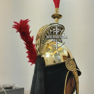 Op de afbeelding: Een goudkleurige helm met een rode pluim en zwarte details. De helm heeft ingewikkelde patronen en een zwarte, vloeiende staart. De tekst "MEDIEVAL RELIC" is zichtbaar op de helm.