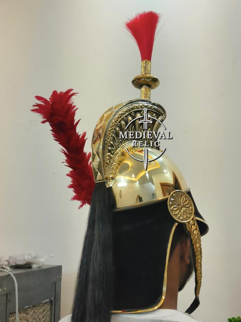Op de afbeelding: Een goudkleurige helm met een rode pluim en zwarte details. De helm heeft ingewikkelde patronen en een zwarte, vloeiende staart. De tekst "MEDIEVAL RELIC" is zichtbaar op de helm.
