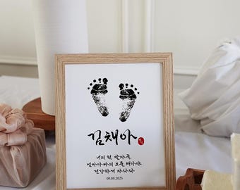 May include: A framed print with a light wood frame features two black footprints above Korean text. The text includes a name and a date, 09.08.2025. A white vase and decorative items are in the background.
