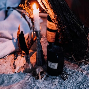 Może przedstawiać: Biała świeca w szklanym świeczniku, czarna butelka z etykietą z napisem "AMBRE", szklana butelka, czaszka i kawałek poroża na piaszczystym podłożu.