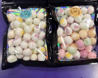 May include: Two black, resealable bags filled with colorful, round candies. The candies are white with pastel-colored centers and are labeled with a circular sticker that reads "RY'S FREEZE DRIED CANDY". The bags sit on a purple surface.