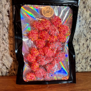 Può includere: Un sacchetto nero richiudibile pieno di caramelle liofilizzate. Le caramelle sono di un rosso brillante con zuccherini colorati. Il sacchetto ha una finestra trasparente e iridescente. È visibile un adesivo circolare con la scritta "RY'S FREEZE DRIED CANDY".