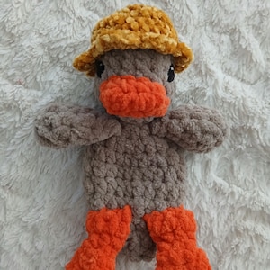 May include: A handmade crochet duck toy with a brown body, orange beak and feet, and a yellow hat. The plush duck is made of soft yarn and has black eyes. The toy is designed to be a cute and cuddly item.