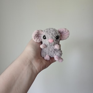May include: A small, handmade crochet mouse plush toy crafted from soft, textured grey yarn. The mouse features large, rounded ears with light pink inner details, a small pink nose, and two shiny black plastic safety eyes. It has tiny pink paws.