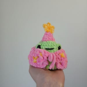 Op de afbeelding: Een kleine groene gehaakte kikker die een roze en gele gehaakte hoed draagt met een ster erop. De kikker houdt een roze en gele gehaakte bloem in zijn hand.