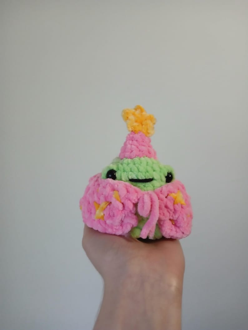 Op de afbeelding: Een kleine groene gehaakte kikker die een roze en gele gehaakte hoed draagt met een ster erop. De kikker houdt een roze en gele gehaakte bloem in zijn hand.
