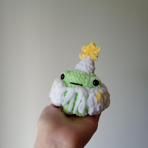 Op de afbeelding: Een groene gehaakte kikker die een witte gehaakte bloem draagt met gele accenten en een gele ster bovenop.