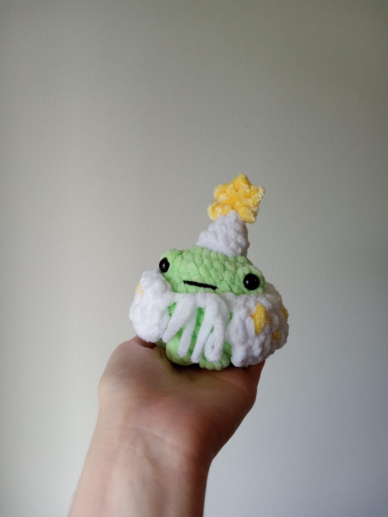 Op de afbeelding: Een groene gehaakte kikker die een witte gehaakte bloem draagt met gele accenten en een gele ster bovenop.