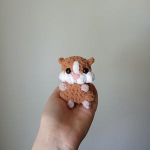 May include: A small, handmade crochet hamster plush toy made from soft, fuzzy brown yarn. It features a white patch on its face, a small pink nose, two black plastic eyes, and tiny light pink paws. The texture is plush and rounded in shape.