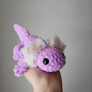 May include: A handmade, plush axolotl in a soft lavender color. The amphibian has fluffy, light pink gills and black safety eyes. The stuffed animal is made of a textured yarn and is held in a hand.