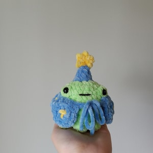 Op de afbeelding: Een klein groen gehaakt pluche speelgoedje dat een blauwe gehaakte hoed draagt met gele sterren en een gele ster bovenop. Het speelgoed heeft zwarte ogen en een kleine mond.