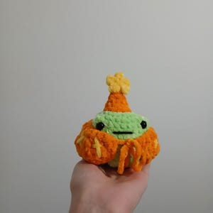 Op de afbeelding: Een groen gebreid pluche speelgoed dat een oranje en gele gebreide hoed en sjaal draagt. Het speelgoed heeft zwarte ogen en een kleine mond.