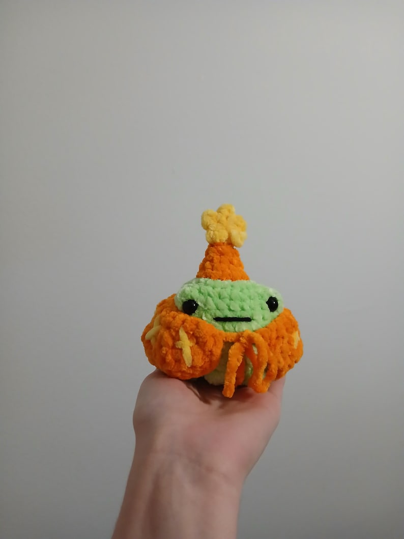Op de afbeelding: Een groen gebreid pluche speelgoed dat een oranje en gele gebreide hoed en sjaal draagt. Het speelgoed heeft zwarte ogen en een kleine mond.