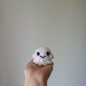 May include: A small, white crocheted ghost with a smiling face and rosy cheeks. The handmade amigurumi figure has black eyes and a stitched mouth. The ghost is held in a hand against a plain background.