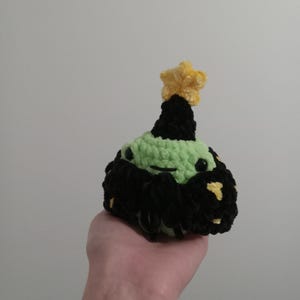 Op de afbeelding: Een groen gehaakt pluche speelgoed met een zwarte en gele stervormige hoed. Het speelgoed heeft een zwarte gehaakte strik en gele stippen op zijn lichaam.