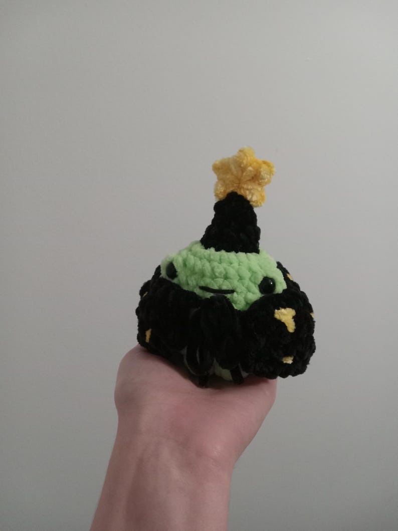 Op de afbeelding: Een groen gehaakt pluche speelgoed met een zwarte en gele stervormige hoed. Het speelgoed heeft een zwarte gehaakte strik en gele stippen op zijn lichaam.