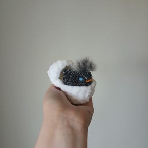May include: A handmade, crocheted baby bird nestled in a white, fluffy crocheted egg. The bird is primarily black with a gray feather and blue and orange accents. The egg is held in a hand against a plain background.