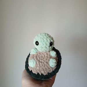 May include: A handmade plush turtle in shades of mint green, brown, and dark green. The turtle has a round head, small black eyes, and a shell-like body. The plush toy is soft and cuddly, perfect for children or as a decorative item.