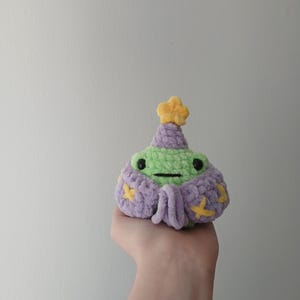 Op de afbeelding: Een groene gehaakte kikker die een paarse en gele gehaakte hoed en een gewaad draagt. De kikker houdt een kleine gele ster in zijn hand.