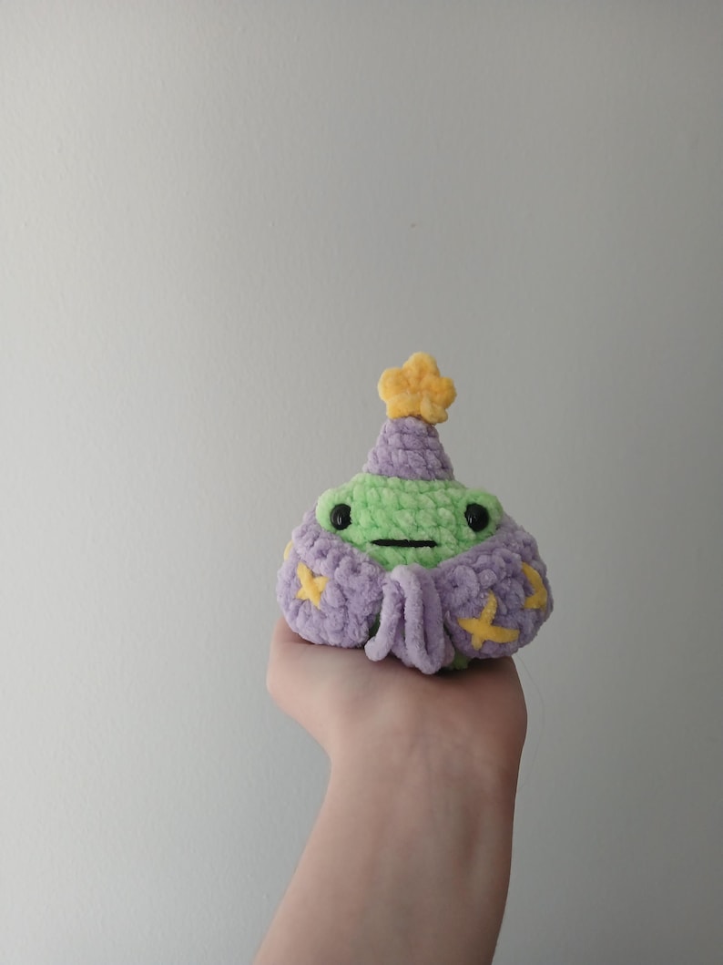 Op de afbeelding: Een groene gehaakte kikker die een paarse en gele gehaakte hoed en een gewaad draagt. De kikker houdt een kleine gele ster in zijn hand.