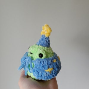 Op de afbeelding: Een groene gehaakte kikker met een blauwe en gele feestmuts. De kikker houdt een blauwe en gele gehaakte sjaal vast.
