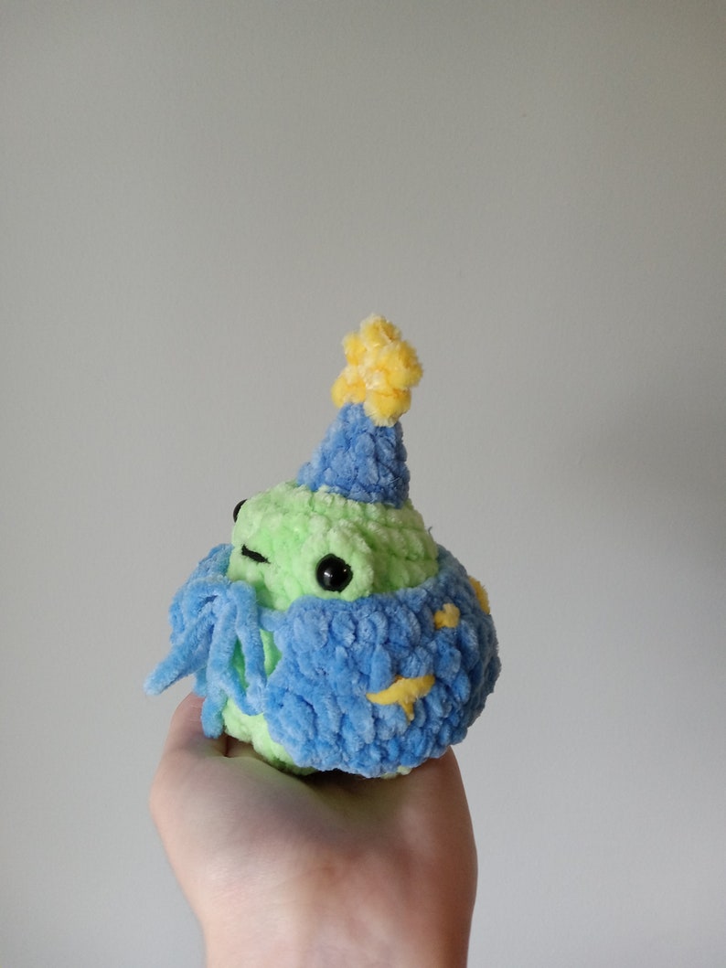 Op de afbeelding: Een groene gehaakte kikker met een blauwe en gele feestmuts. De kikker houdt een blauwe en gele gehaakte sjaal vast.
