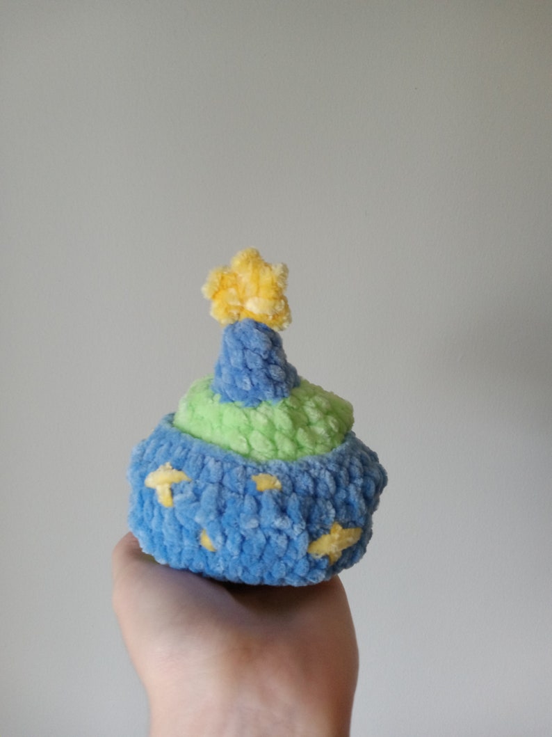 Op de afbeelding: Een blauw en groen gebreid pluche speelgoed in de vorm van een cupcake met een gele ster bovenop.