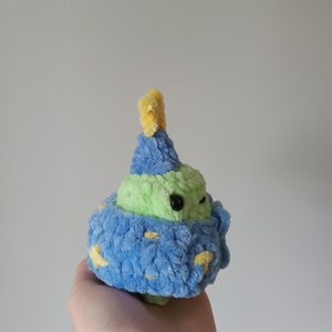 Op de afbeelding: Een groen en blauw gehaakt pluche speelgoed met een gele hoed. Het speelgoed heeft een klein, zwart oog en een blauw en geel gepatroneerd lichaam.