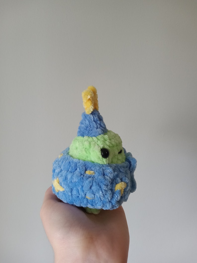 Op de afbeelding: Een groen en blauw gehaakt pluche speelgoed met een gele hoed. Het speelgoed heeft een klein, zwart oog en een blauw en geel gepatroneerd lichaam.